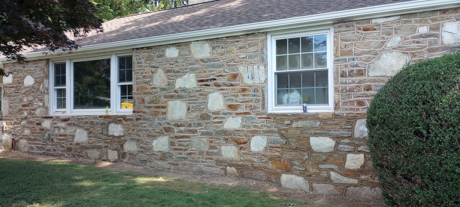 Stone exterior wall of a house with two windows, grass in front, and a trimmed bush on the right.