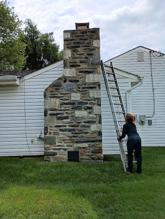 Stone chimney beside a house, with a woman standing on a ladder next to it.