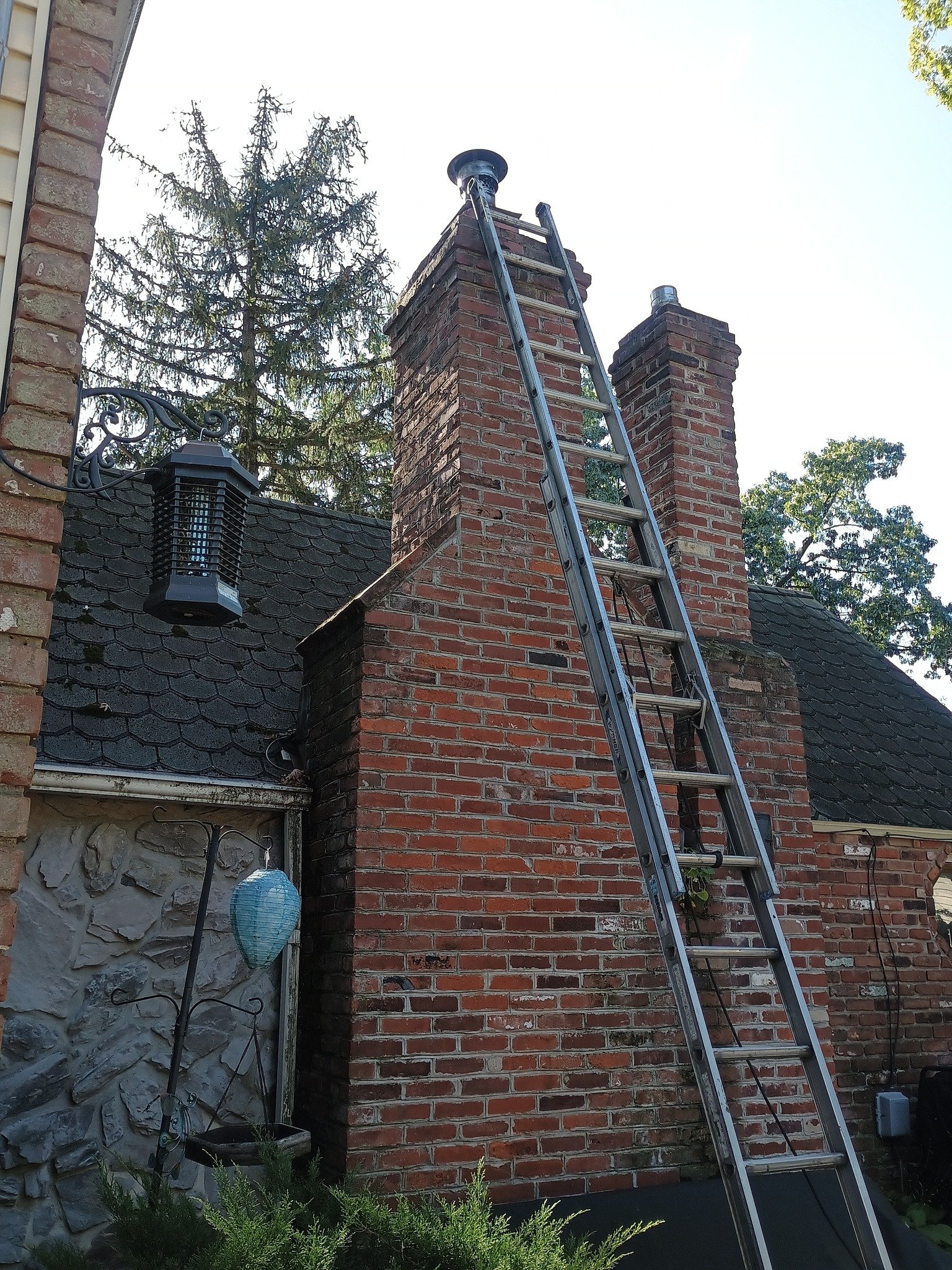 Aluminum ladder leaning against a brick chimney on a house roof, with trees and a bright sky behind it.