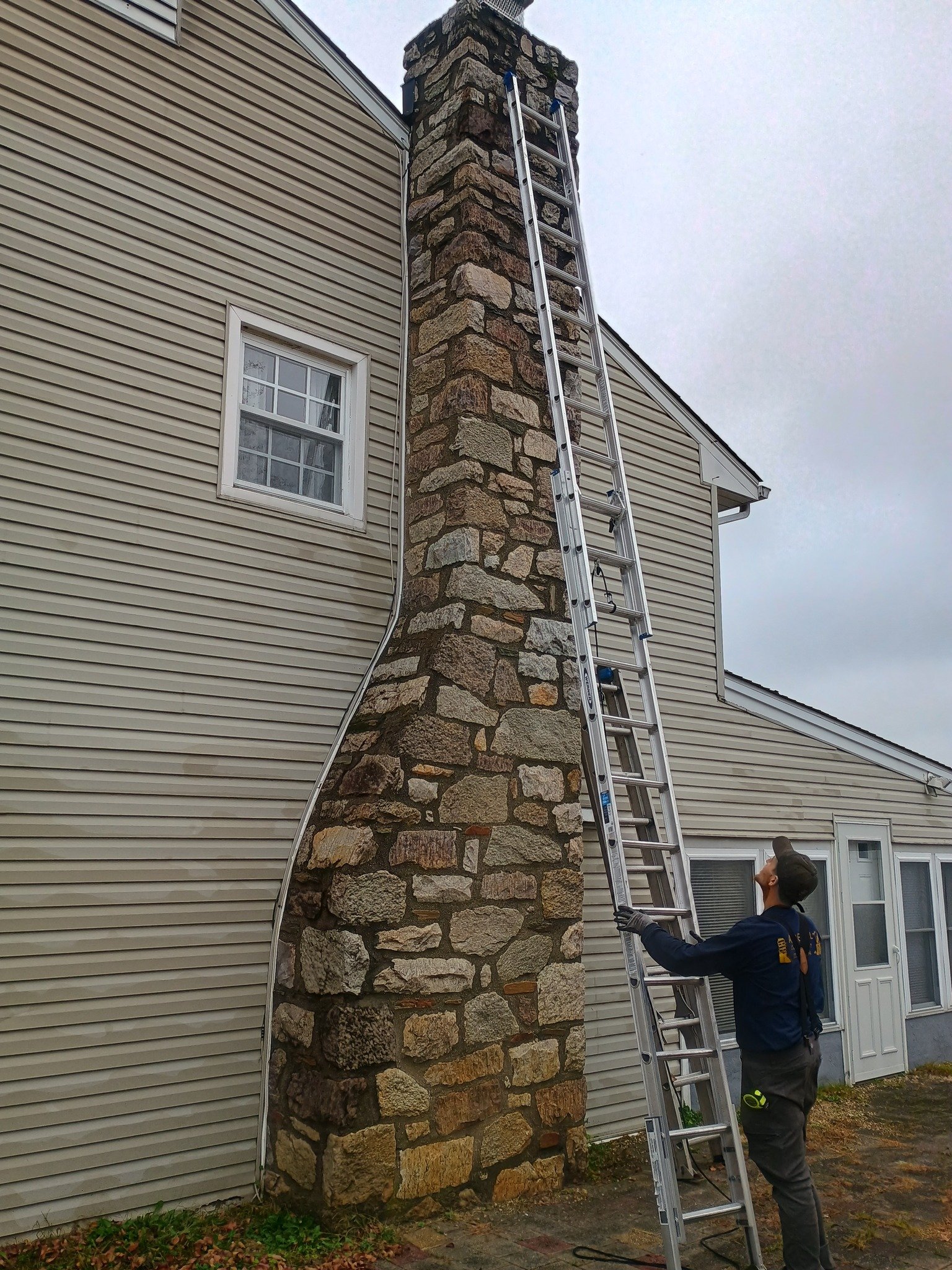 Team member holding a ladder beside a tall stone chimney on the side of a house