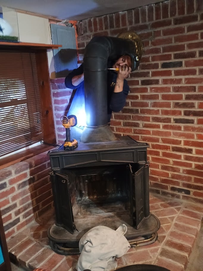 Team member working on a black wood stove in a brick room, using a drill and flashlight.