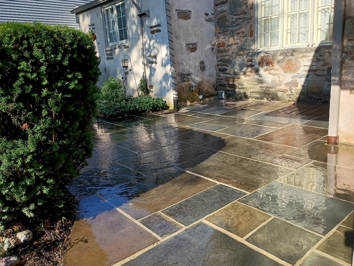 Wet stone patio beside a house with a trimmed hedge and sunlit stone wall